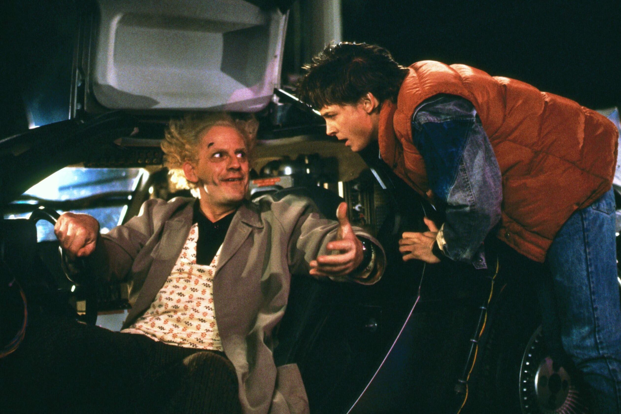 Scientist gestures excitedly inside a futuristic car, speaking to an attentive young man leaning in, set in a dimly lit environment.
