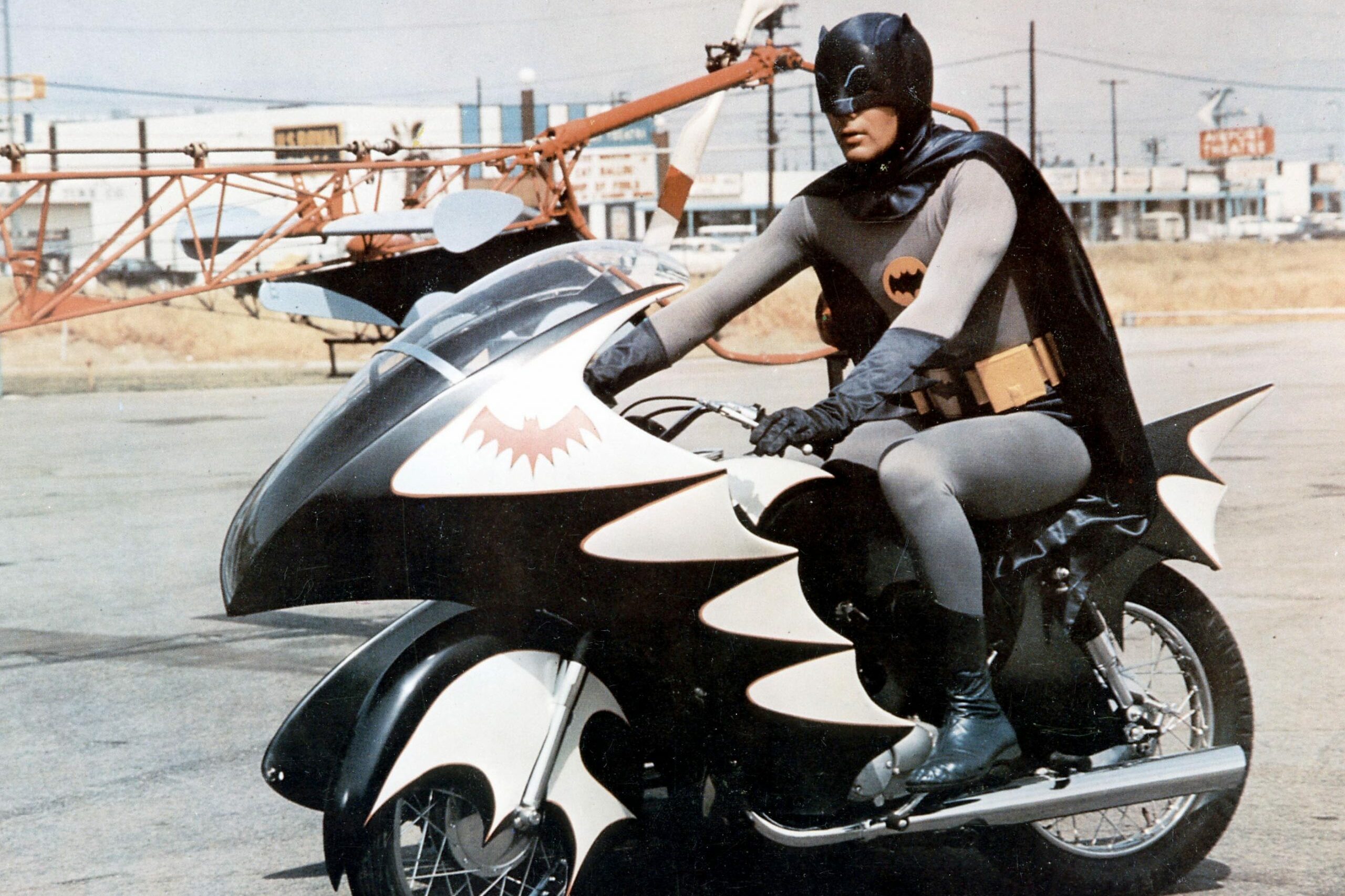 A person in a bat-themed costume rides a custom motorcycle shaped like a bat, parked in an industrial area.