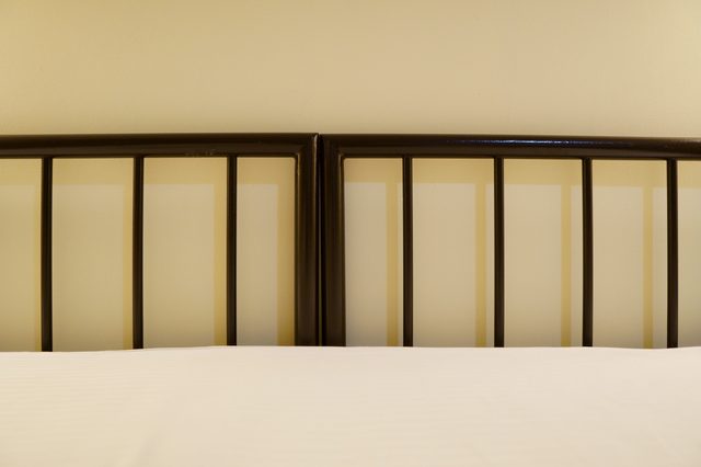 Metal bed frame stands against a beige wall, bordered by a white bedspread below.