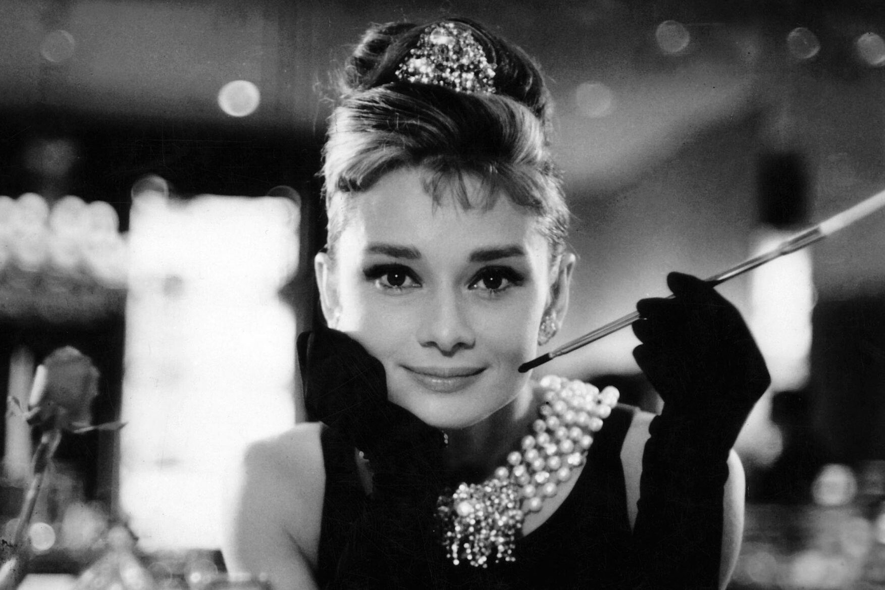 Woman holding cigarette holder, wearing elegant dress and pearl necklace, smiling in a dimly lit, sophisticated setting.