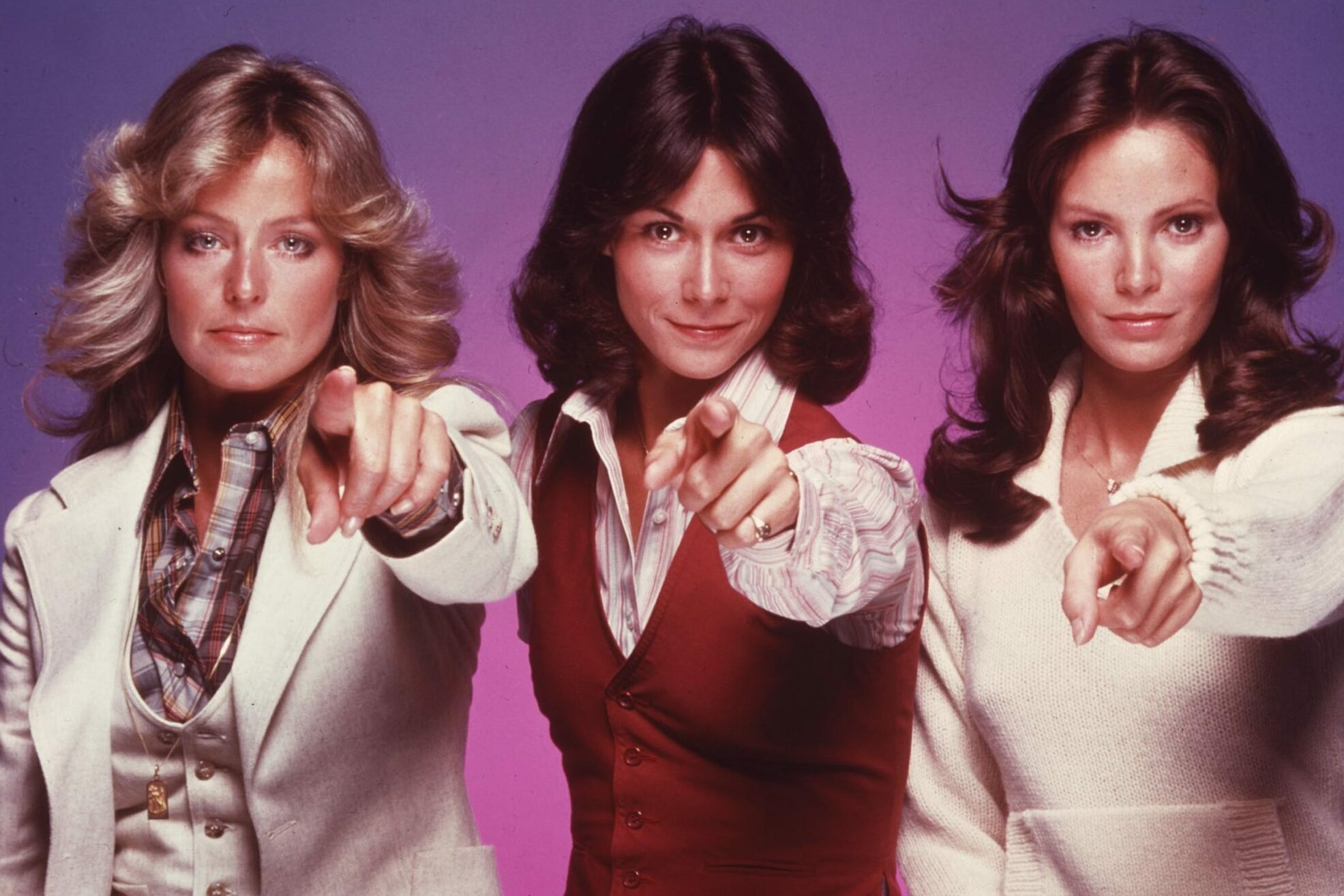 Three women point forward, wearing stylish outfits; one in a white suit, another in a red vest, and the third in a cream sweater.