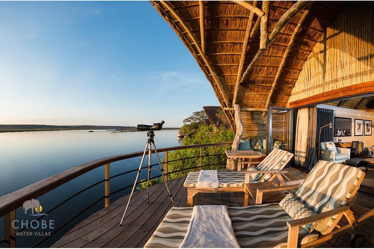 Telescope stands on a wooden deck, overlooking a tranquil river beneath a blue sky; lounge chairs and a thatched villa sit nearby.