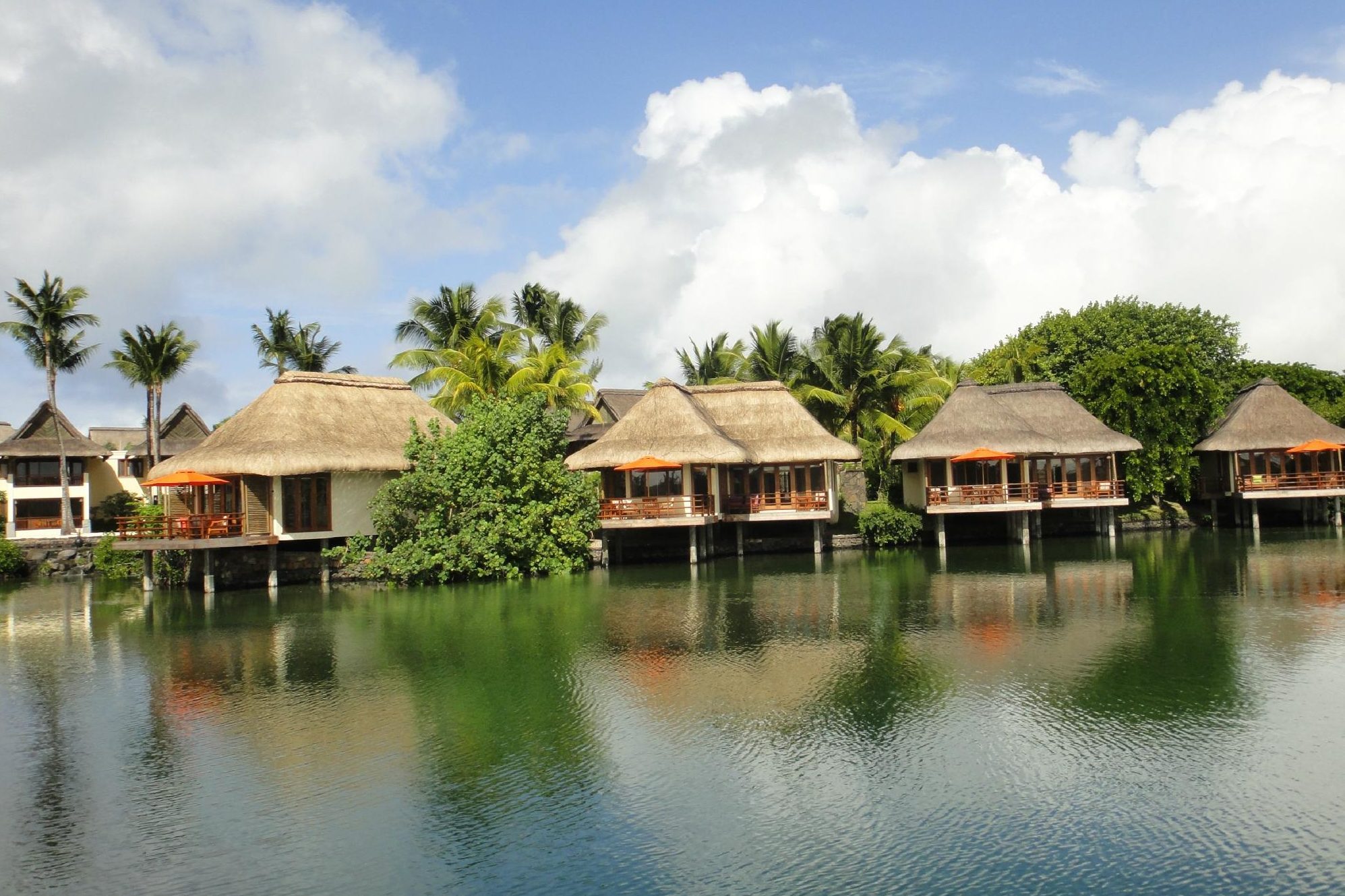 Thatched-roof bungalows rest on stilts over water, surrounded by lush palm trees and greenery, under a bright blue sky with scattered clouds.