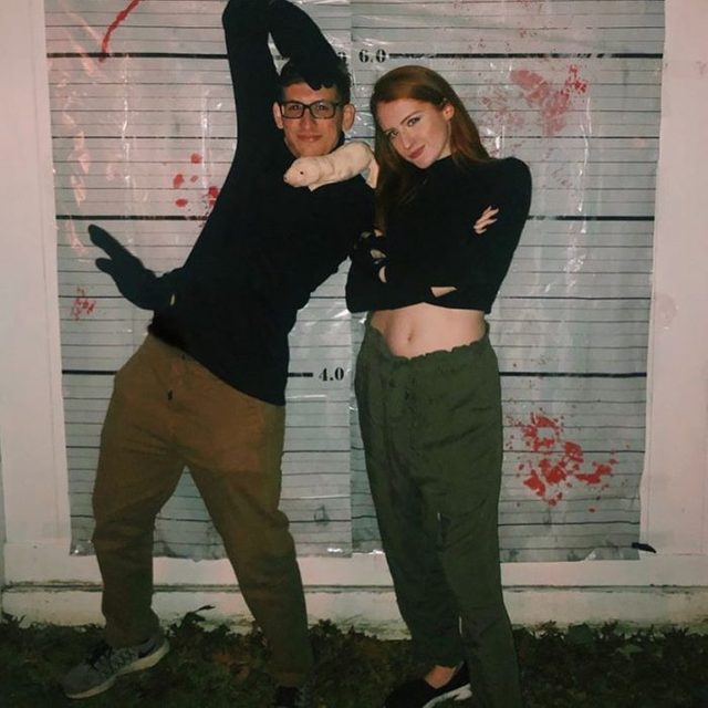 Two people pose playfully, wearing casual clothes, against a height chart backdrop with red splatters, suggesting a crime scene theme.