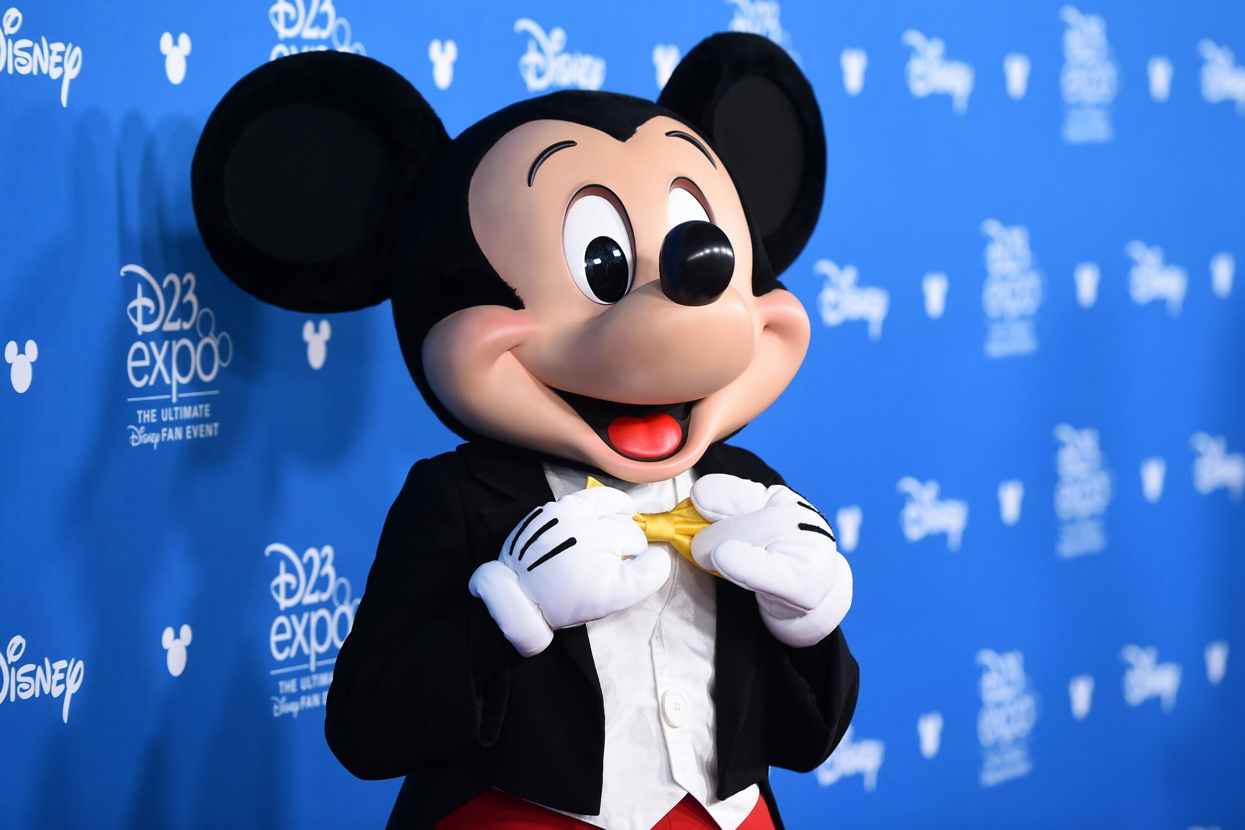 Character holds a bow tie, smiling in front of a blue backdrop with "D23 Expo" and "Disney" logos.