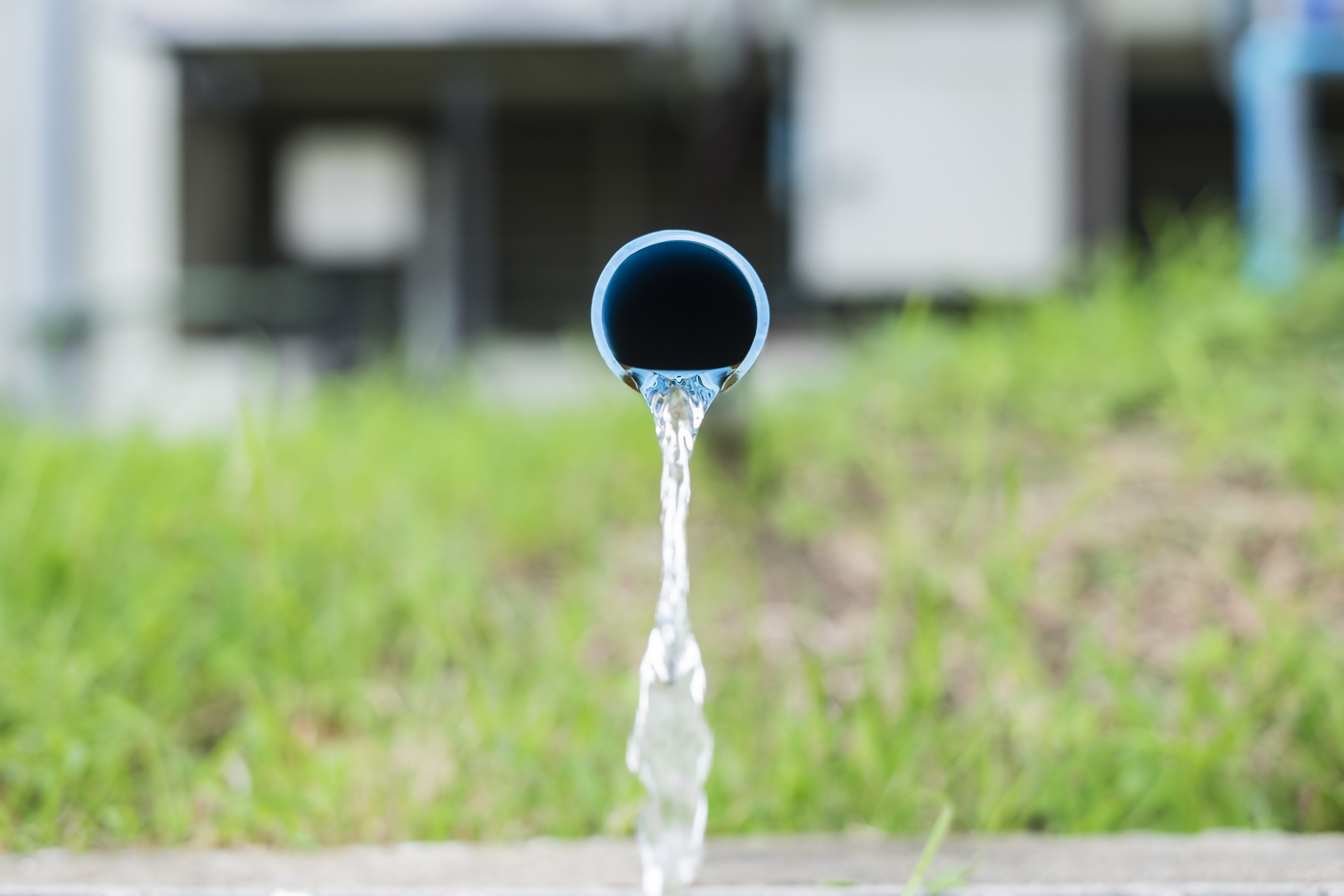 A blue pipe releases flowing water onto grass, with blurred buildings in the background.
