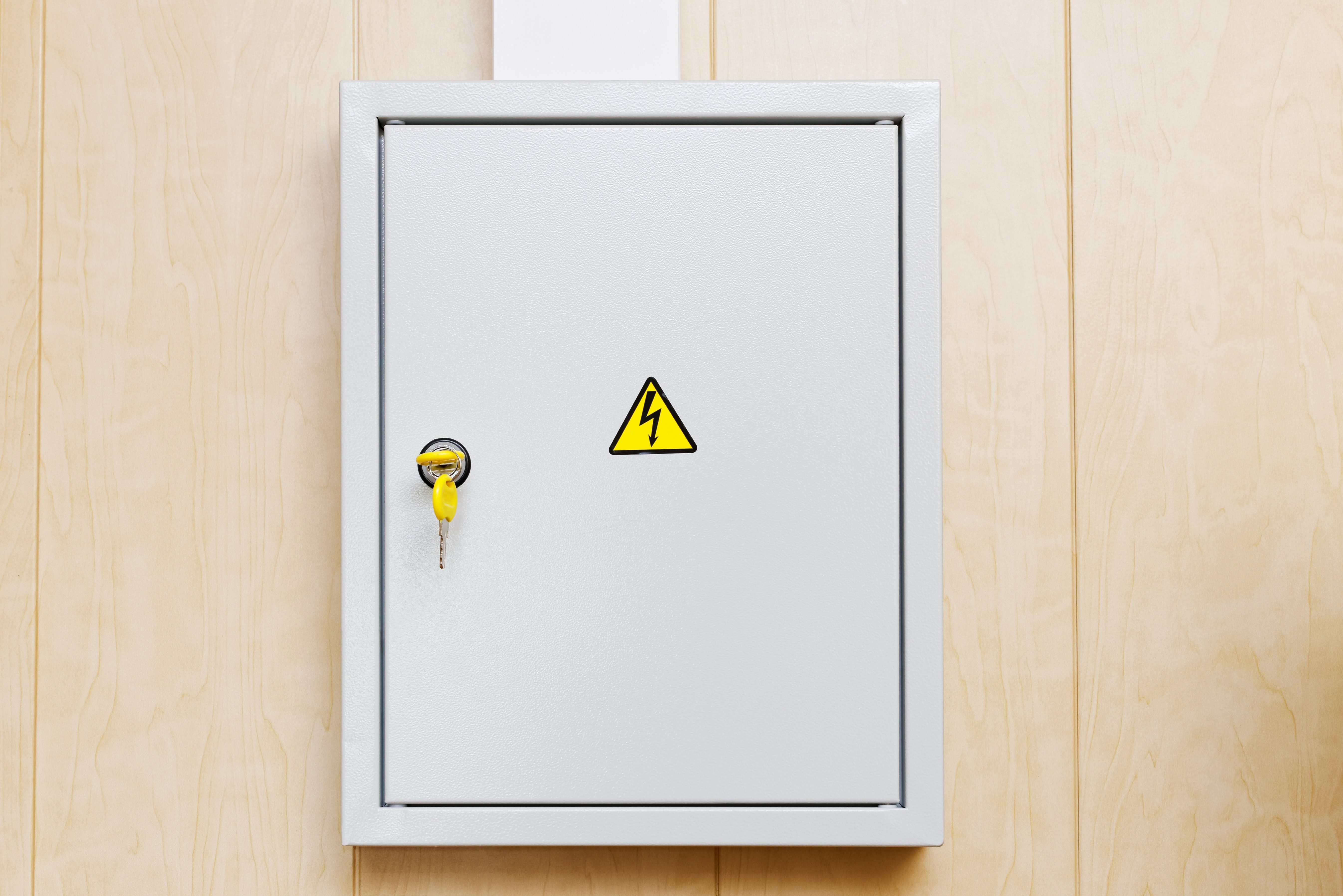 Electrical cabinet with yellow key inserted, mounted on a wooden wall; features a high voltage warning symbol.