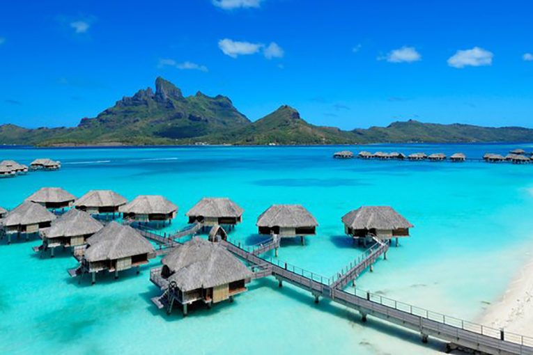 Overwater bungalows sit on turquoise waters, connected by wooden walkways, with lush green mountains under a clear blue sky in the background.