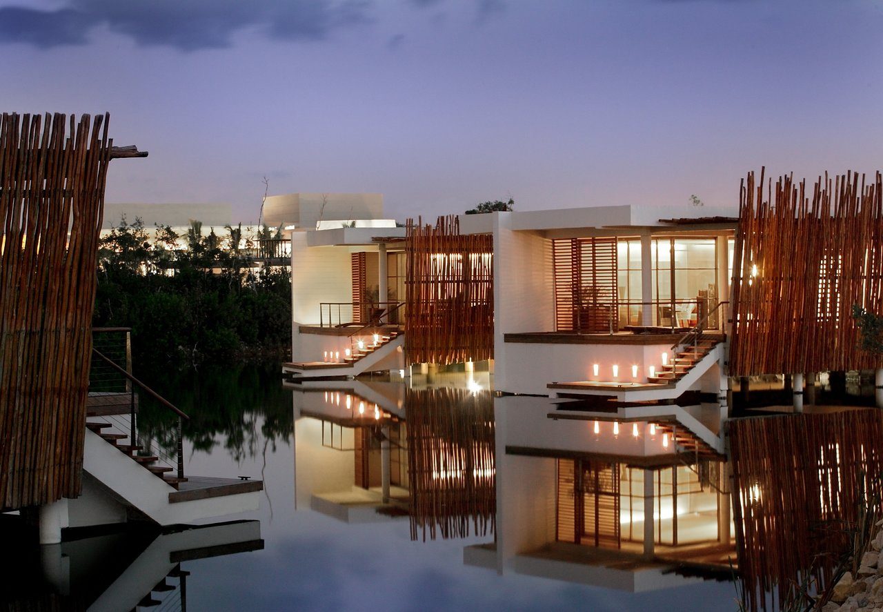 Modern white buildings, surrounded by wooden slats, reflect in calm water under a twilight sky. Warm lights illuminate walkways and interiors.