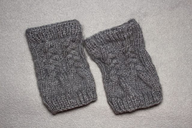 Two gray knitted wrist warmers lie on a textured, light-colored surface.