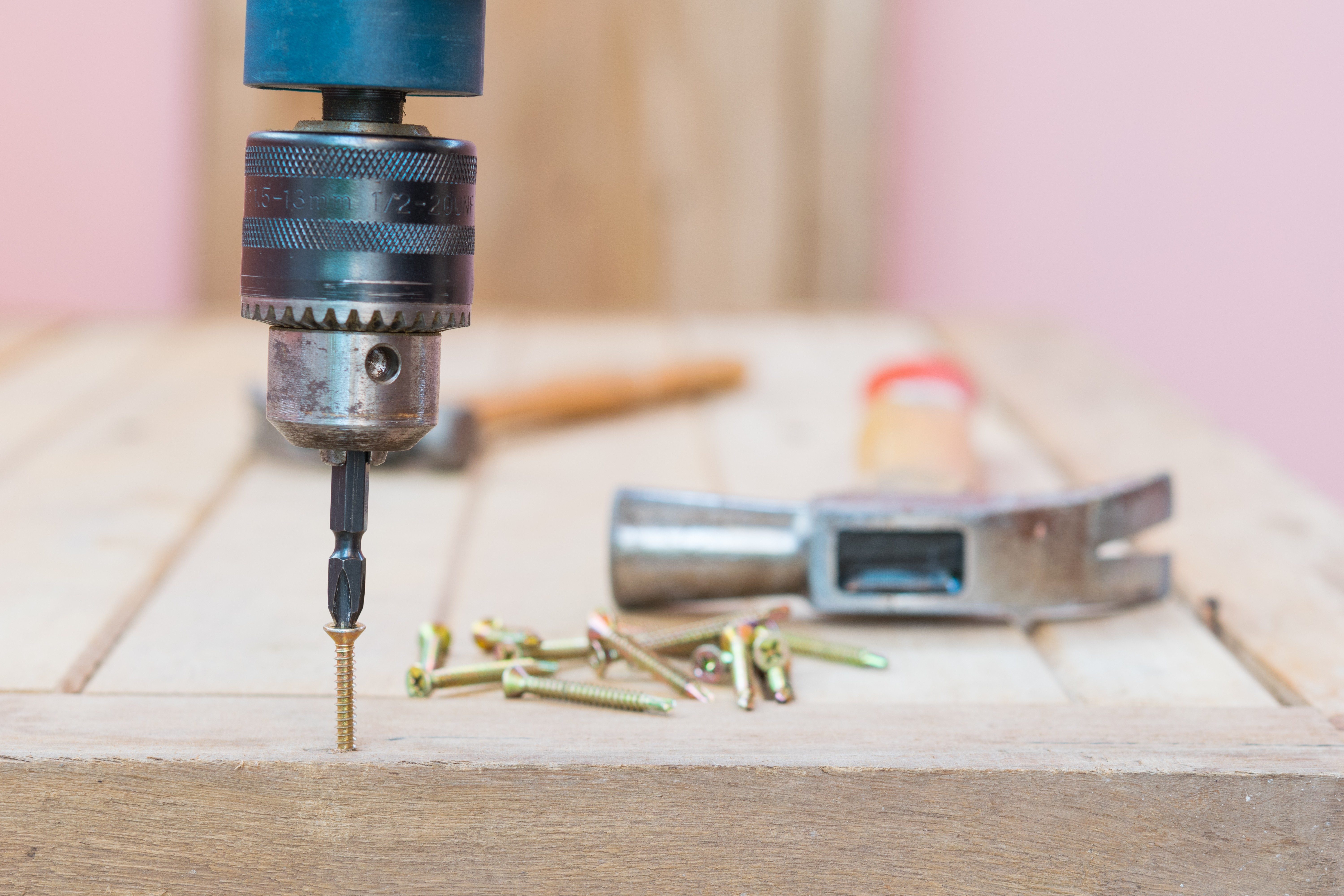 A drill drives a screw into a wooden surface; scattered screws and a hammer lie nearby.