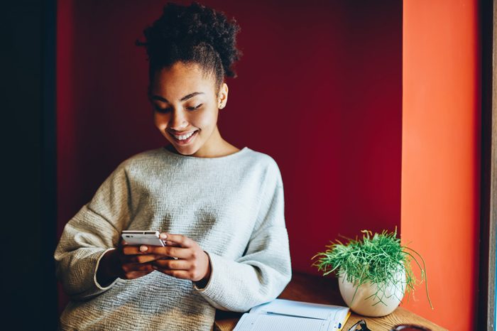 Cheerful dark skinned female blogger checking email and send text message via smartphone on free time, smiling dark skinned hipster girl reading news and updates in social networks using mobile phone