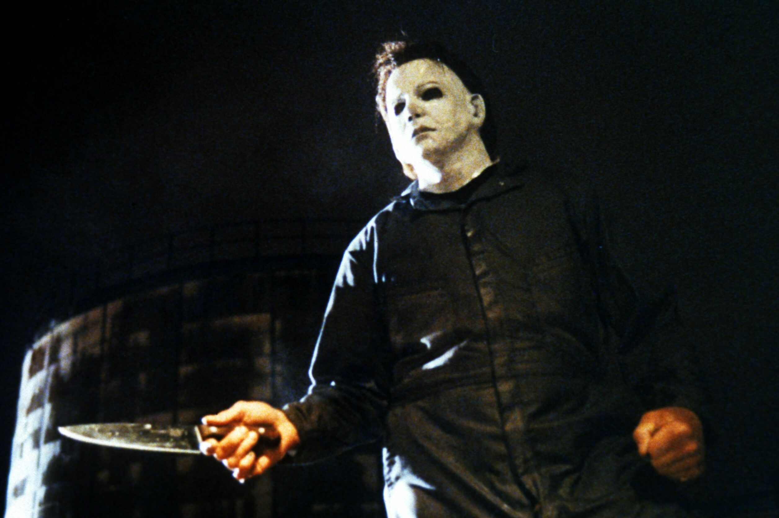 Figure in a white mask holds a knife menacingly, standing in a dark, eerie urban setting with a large building in the background.