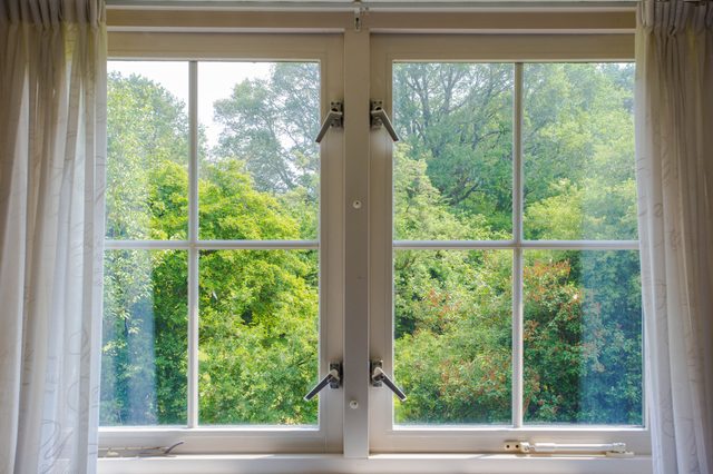 Window opens outward, framed by white curtains, revealing lush green trees under daylight.