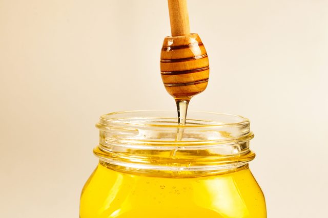 Honey dipper drips golden honey into an open jar against a beige background.