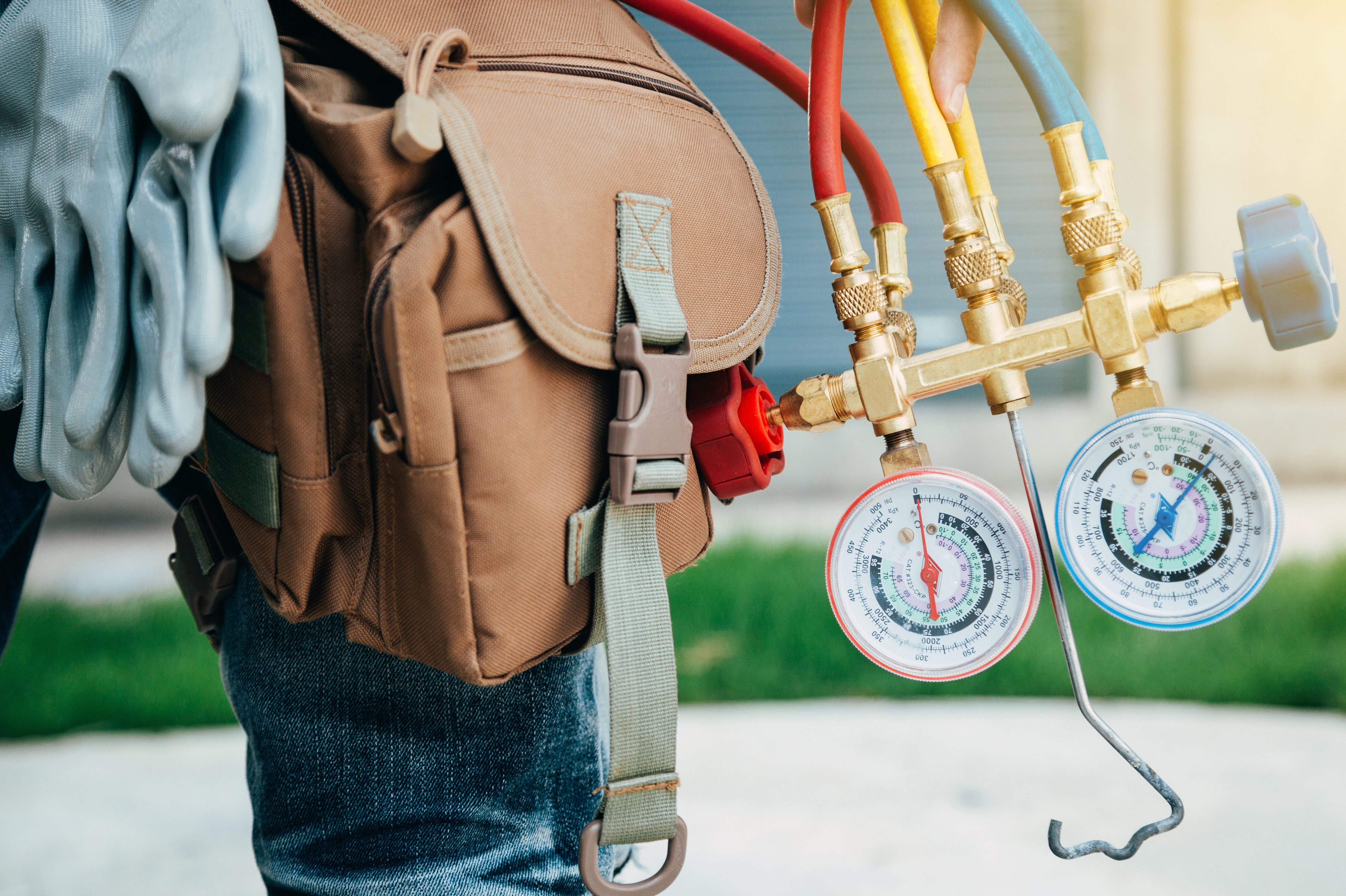 Gauges suspended from hand, connecting hoses behind tools on brown bag and gloves, with pavement and grass in the background.