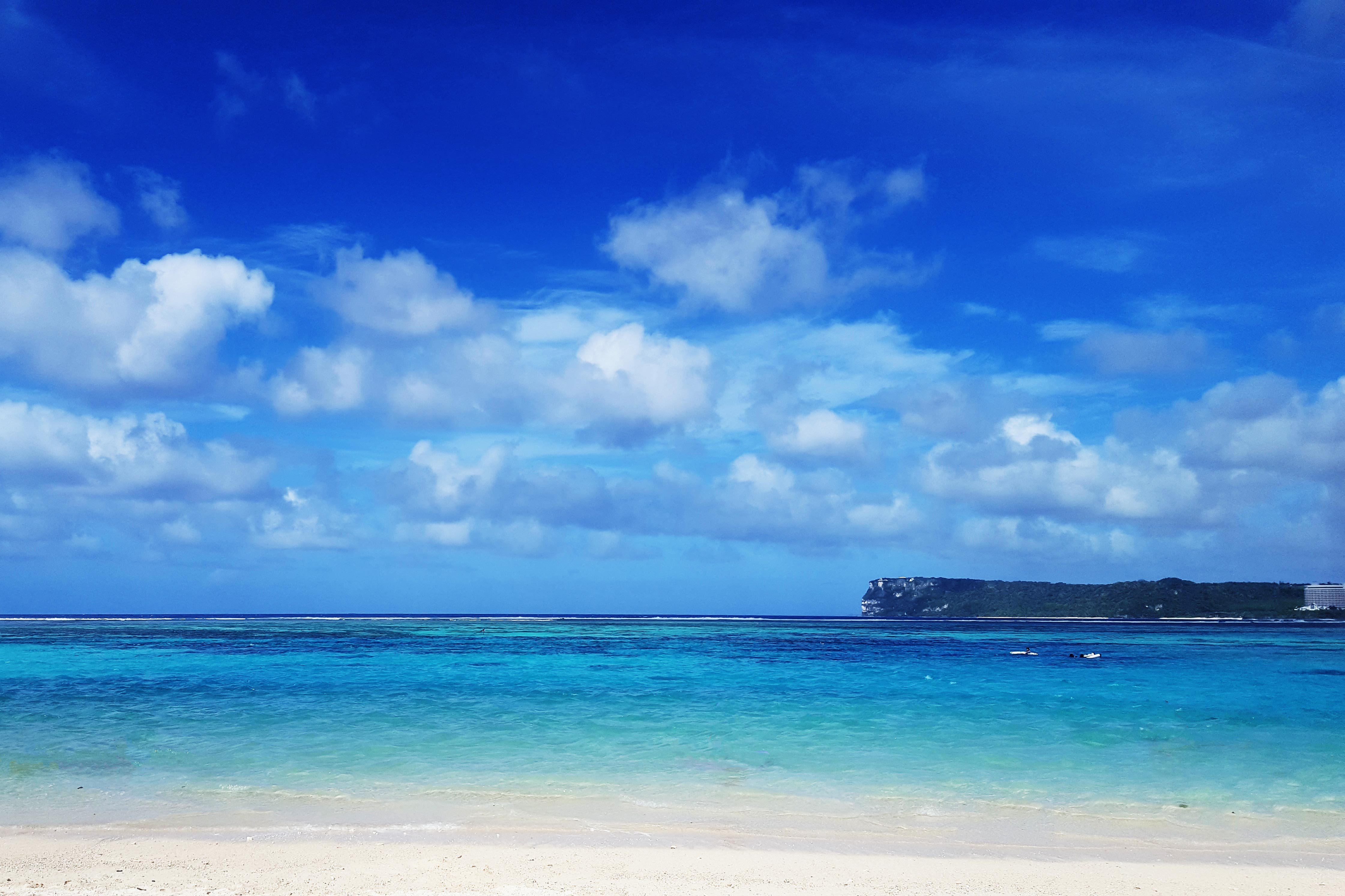 Beach extends into turquoise ocean, bordered by distant green coastline under a bright blue, cloudy sky.