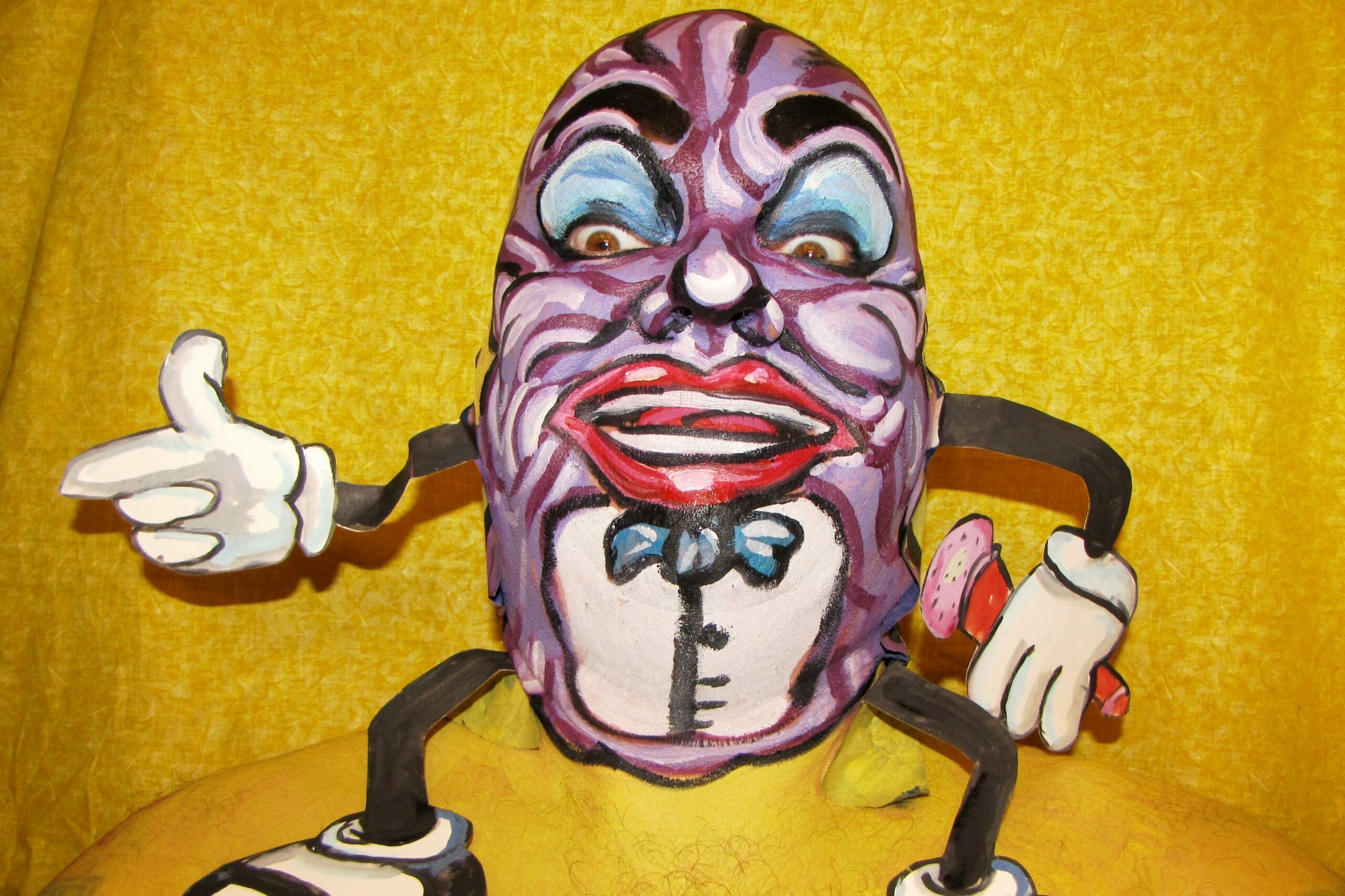 Colorful mask with exaggerated features appears animated, pointing and gesturing with cartoon-like arms, set against a bright yellow background.