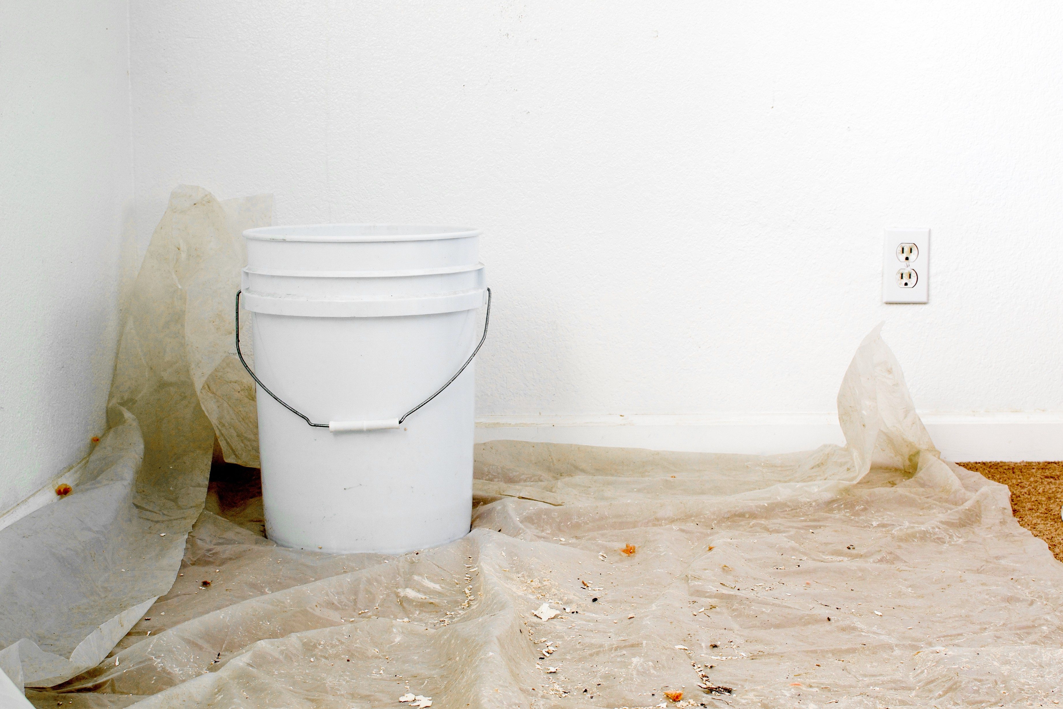 Bucket sits on a dusty, plastic-covered floor in a room with white walls and an electrical outlet.