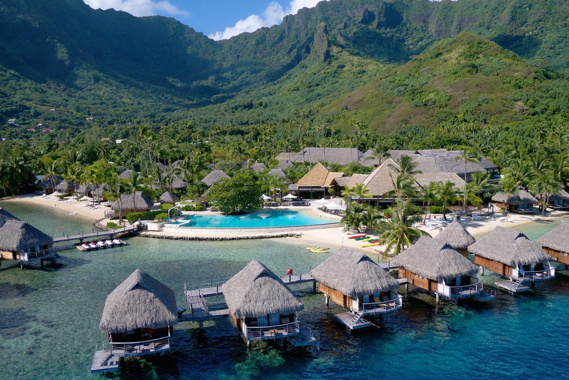 Overwater bungalows sit above clear lagoon; surrounded by lush green mountains and palm trees, adjacent to sandy beach and a large swimming pool.