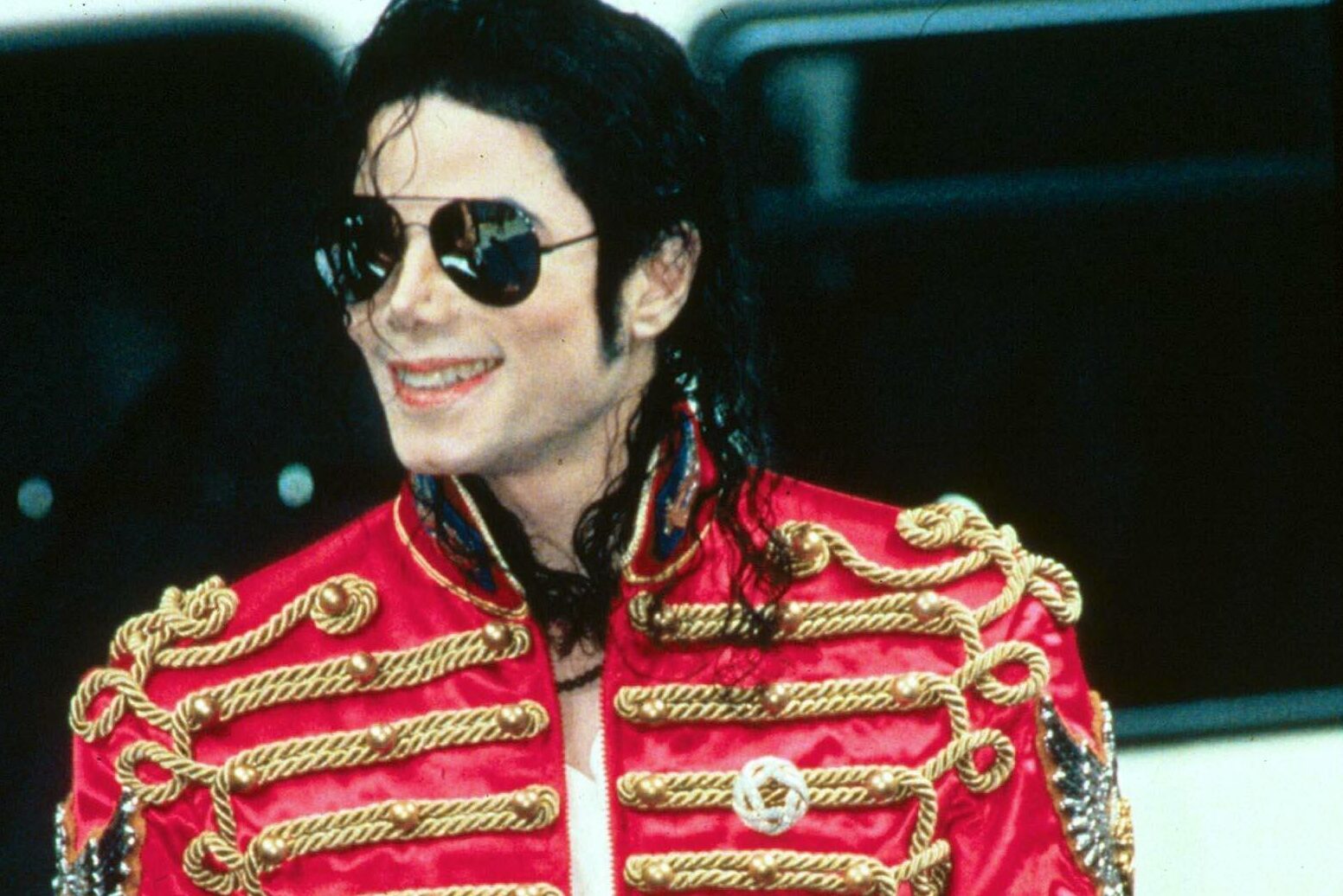 Person wearing a red jacket with gold embellishments smiles, standing outdoors. Sunglasses and dark hair add to the distinctive appearance.