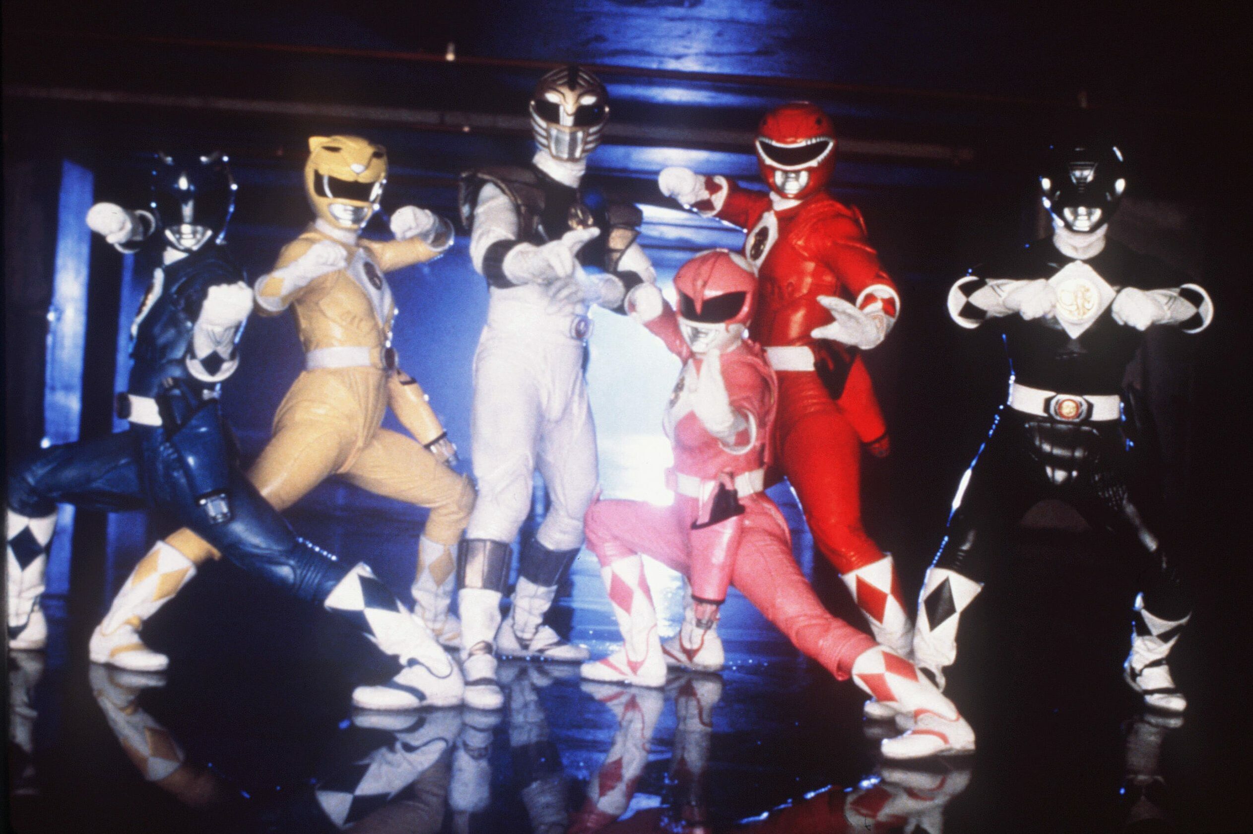 Costumed superheroes strike action poses, displaying dynamic stances in colorful suits. They stand together in a dimly lit environment, suggesting a team formation.