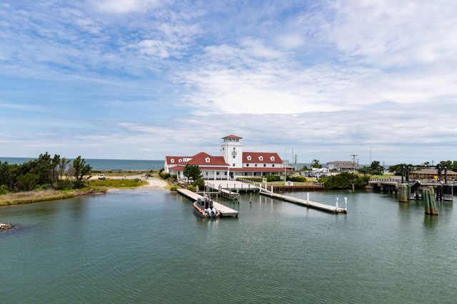 Building with red roofs stands by a waterfront dock, surrounded by small trees, with a distant view of the sea and cloudy sky.
