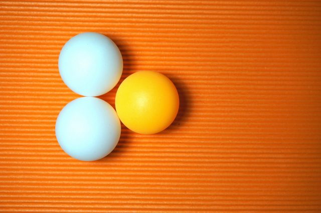 Three ping pong balls rest on an orange striped surface; two are white and one is yellow.