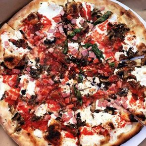 Pizza topped with vegetables, ham, and cheese sits in a brown cardboard box, sliced into pieces with a plastic pizza saver in the center.