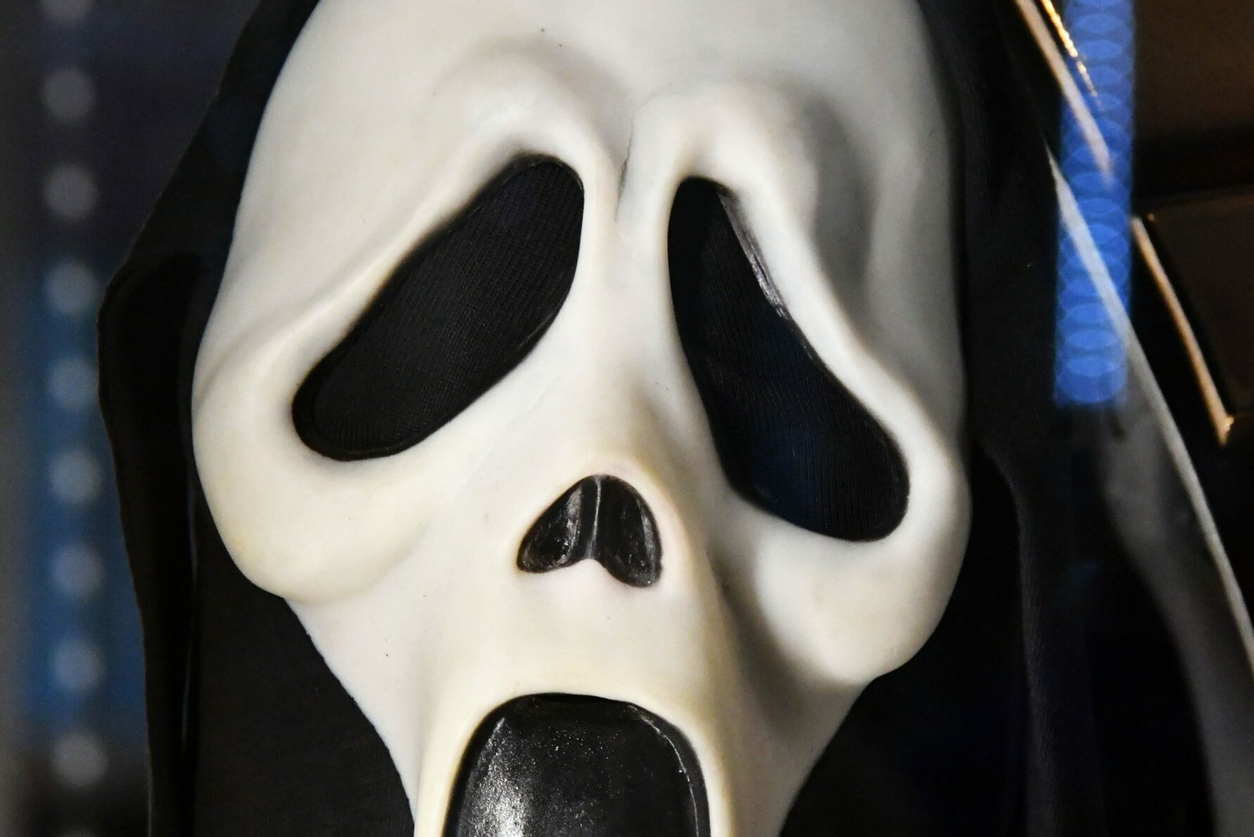 A white, elongated ghost mask with black eye and mouth openings, hangs against a dark background with blue specks.