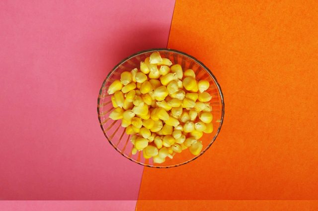 A clear bowl holding yellow corn kernels, resting on pink and orange divided paper background.