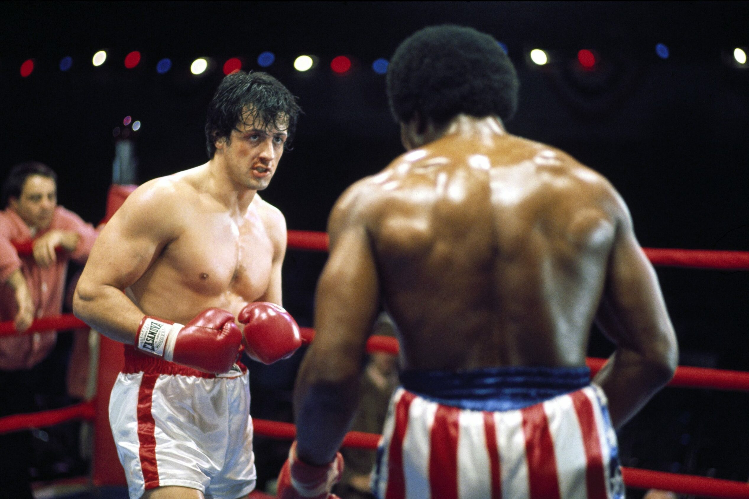 Two boxers face off in a ring, wearing red and white shorts and gloves, under bright colored lights in a crowded arena.