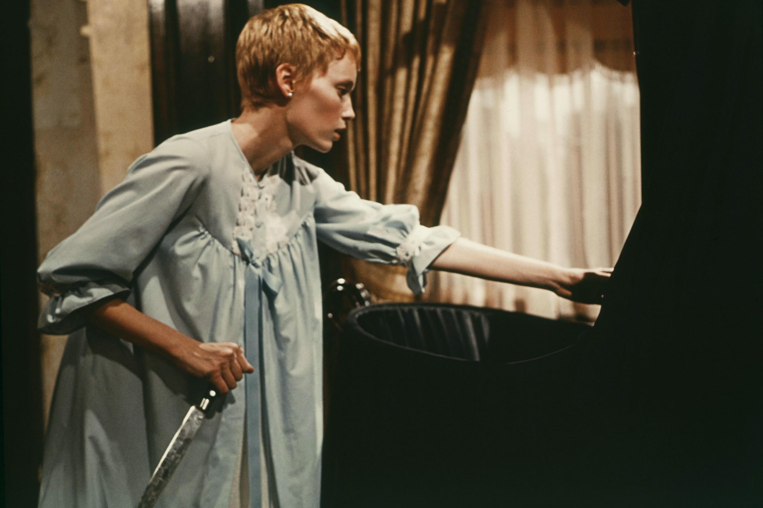 Person in a light blue gown holds a knife, reaching into a black bassinet in a dimly lit room with curtains.