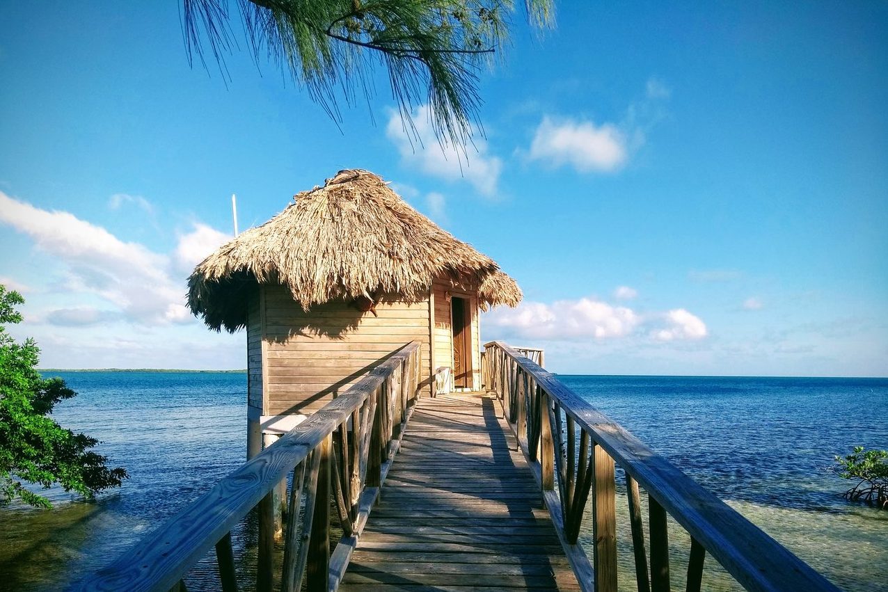 Thatched hut sits on stilts over ocean, connected by wooden walkway; clear blue sky and lush foliage surround.