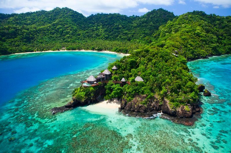 Tropical island showcases huts nestled in lush greenery, encircled by vibrant turquoise waters, under a clear blue sky.