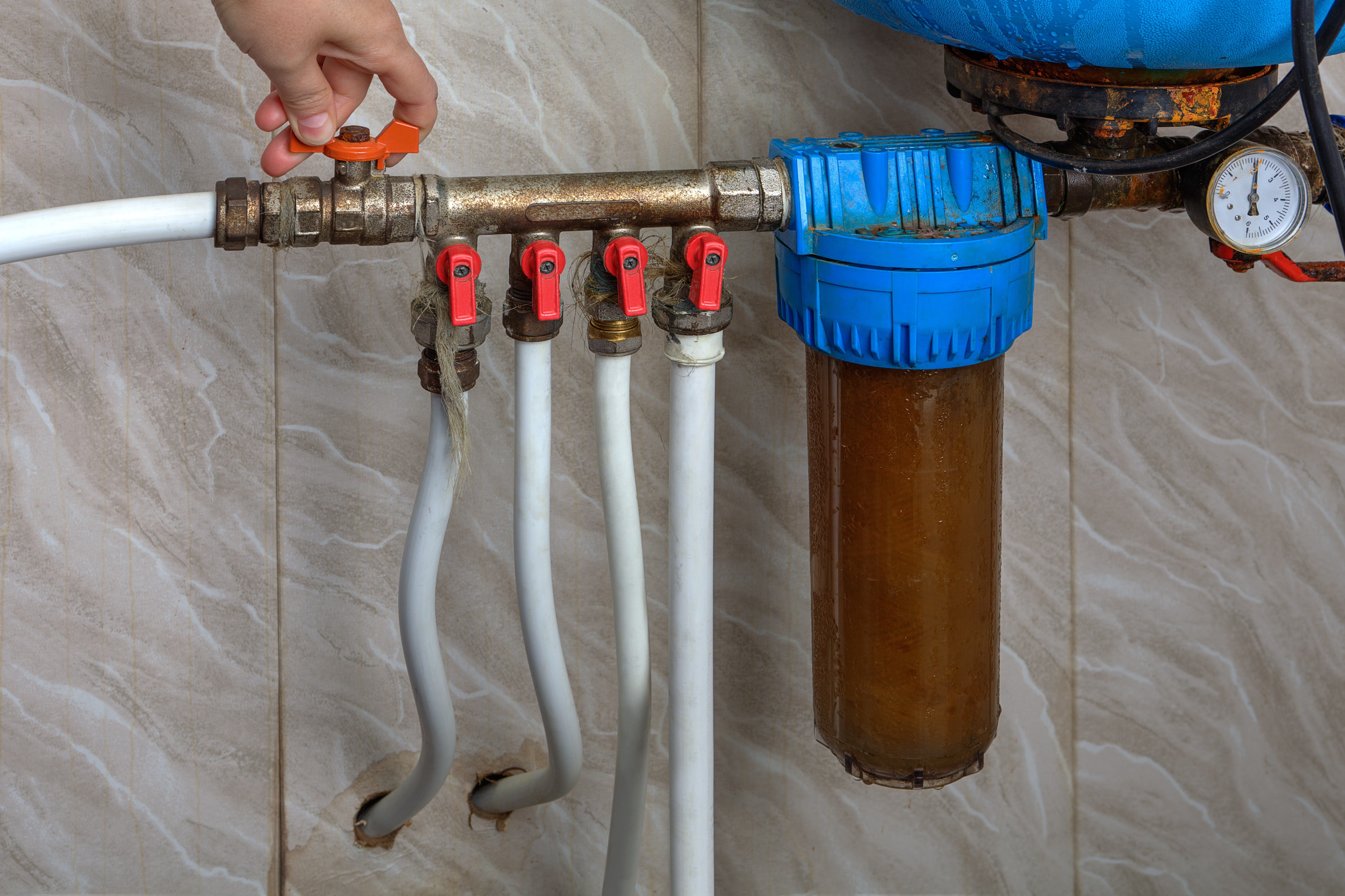 Hand turns valve on a complex plumbing system with several pipes and a blue filter housing, against a tiled wall background.