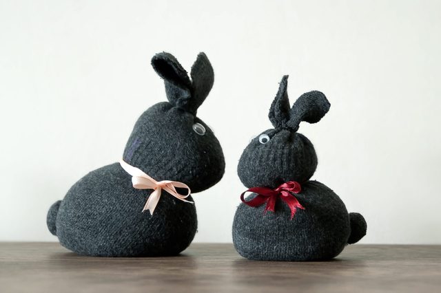 Two sock bunnies sit side by side, adorned with pink and red ribbons, on a wooden surface against a plain background.