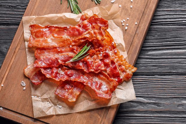 Crispy bacon sits on parchment paper, garnished with rosemary, atop a wooden cutting board on a textured, dark wooden table.