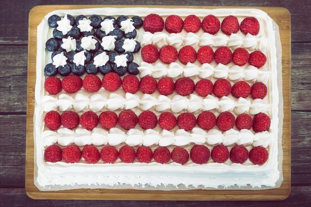 Cake resembling a U.S. flag; blueberries and cream stars, raspberries for stripes, wooden table background.