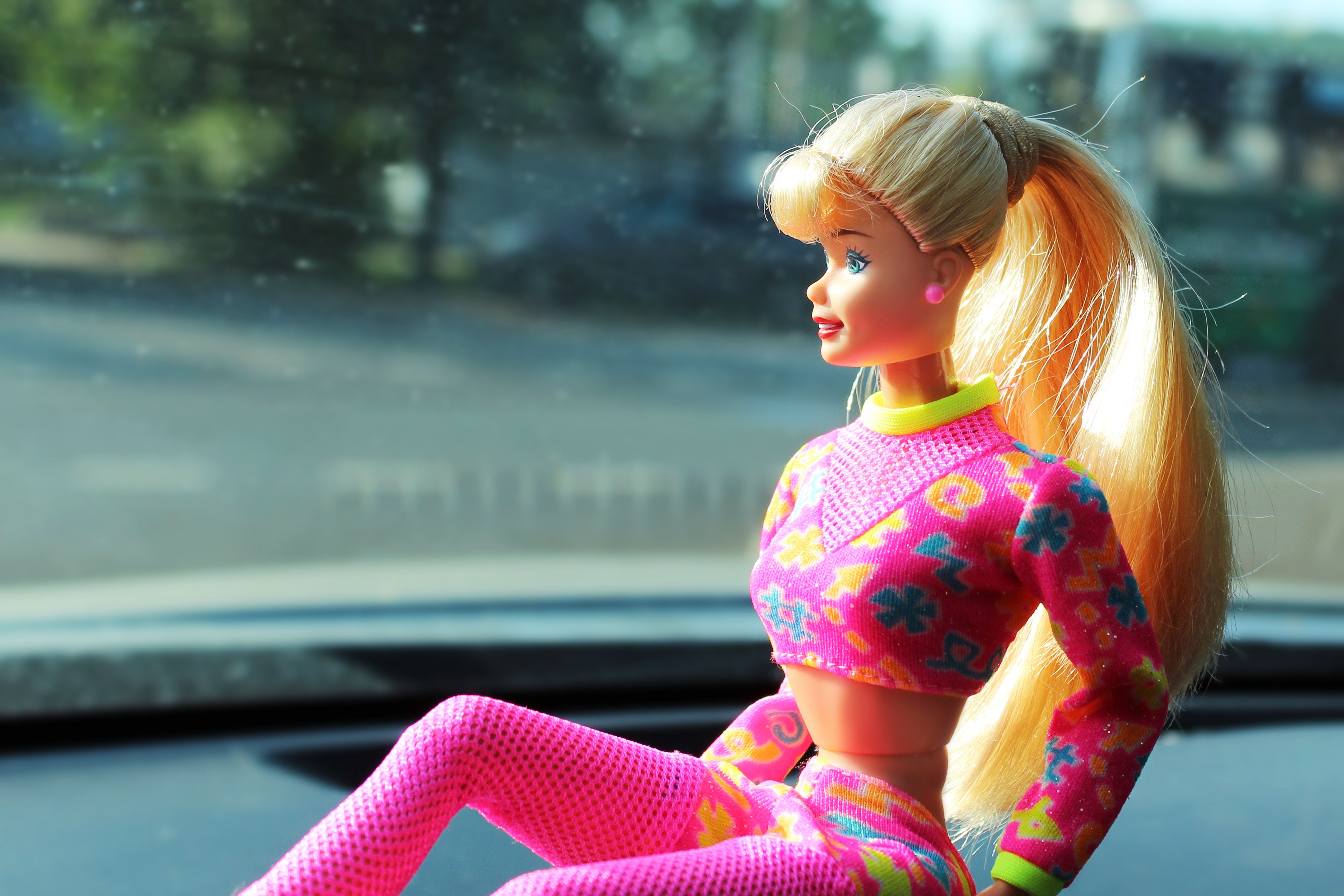 Doll sits on dashboard, wearing colorful floral outfit; background shows a blurred outdoor scene through the windshield.