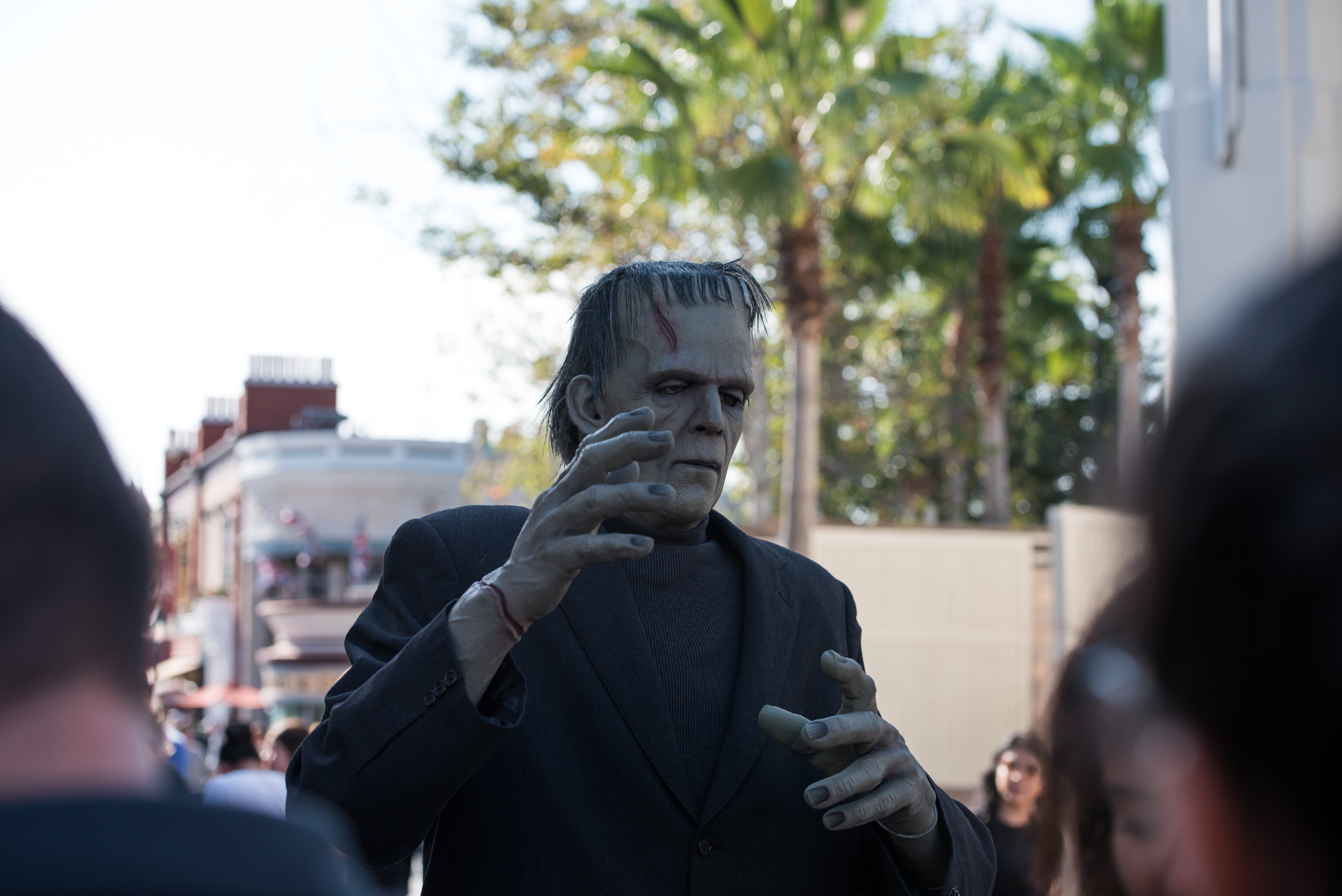 A Frankenstein character raises hands, surrounded by people, in an outdoor setting with palm trees and buildings in the background.