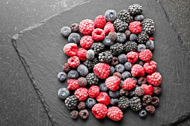 Frozen berries lie scattered on a dark, textured slate surface, showcasing a mix of raspberries, blackberries, and blueberries.