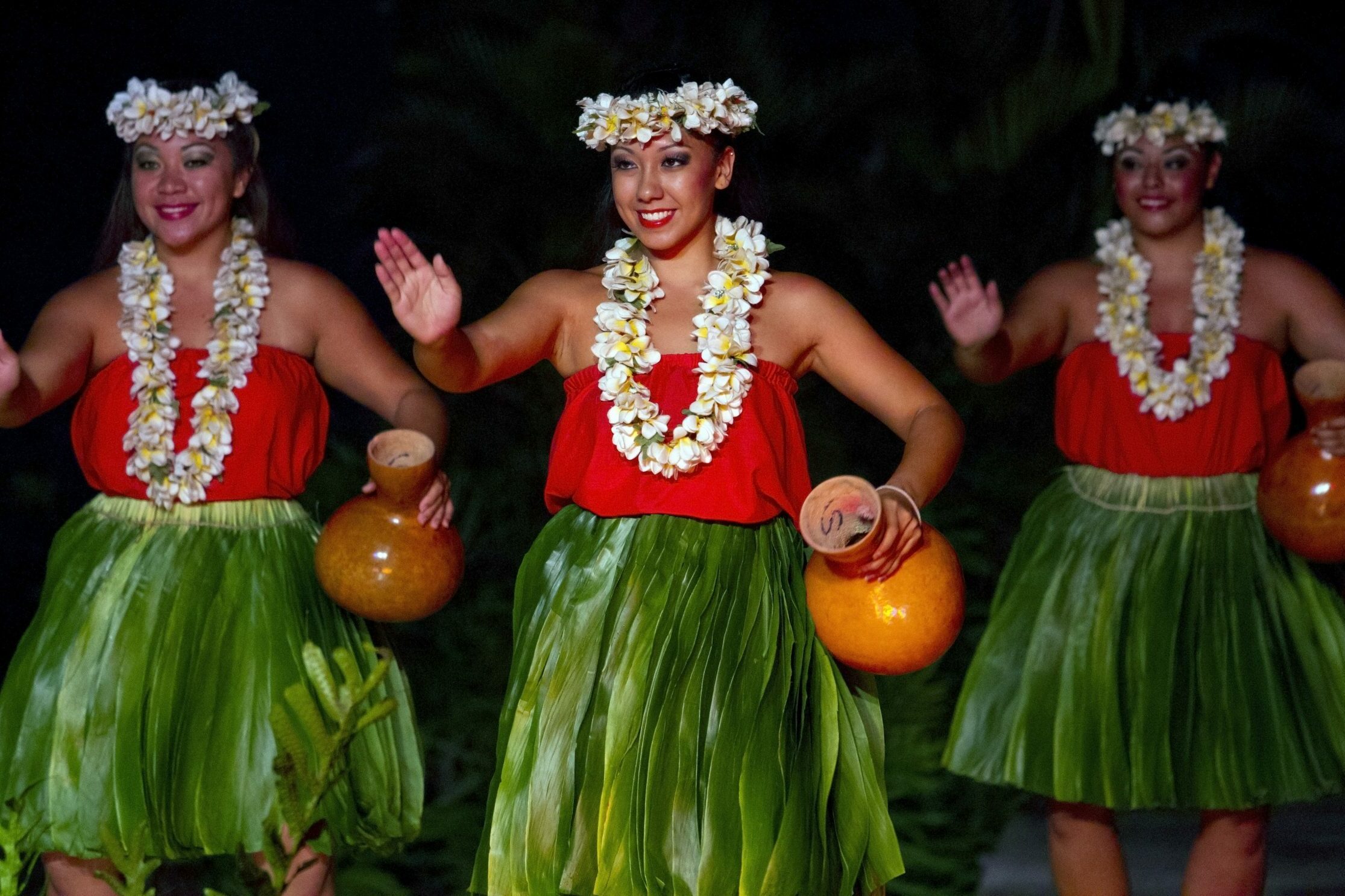 Dancers wearing flower crowns and leis perform with gourds, surrounded by lush greenery. They wear red tops and green hula skirts.