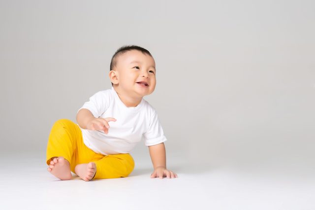 A baby sits and smiles joyfully on a white floor, wearing a white shirt and yellow pants in a bright, neutral space.