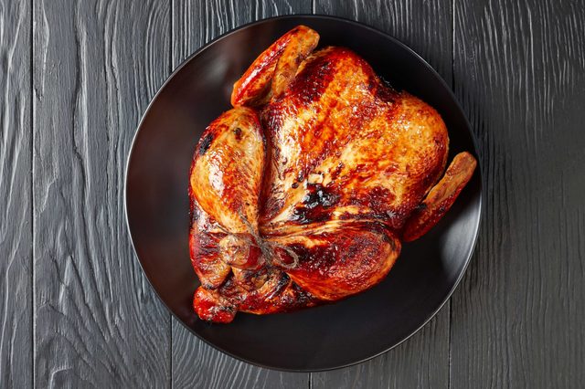 Roasted chicken sits on a black plate, resting atop a textured, dark wooden table.