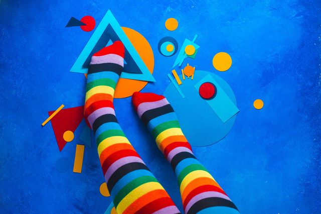 Striped rainbow socks rest on a blue background with colorful geometric paper shapes scattered around.