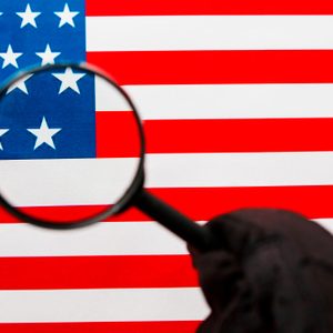 A magnifying glass focuses on stars, held in a gloved hand, against the background of an American flag's red and white stripes.