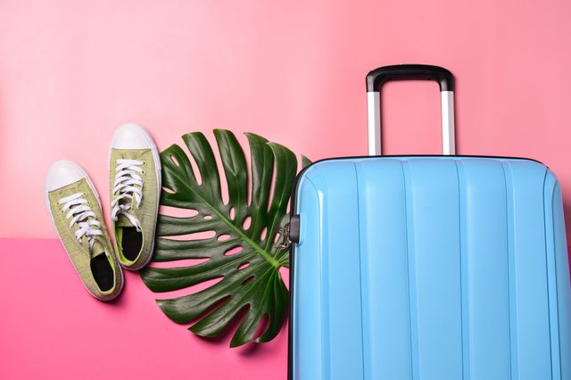 Blue suitcase leans on pink surface; nearby, green sneakers rest beside a large green leaf, suggesting a tropical travel theme.