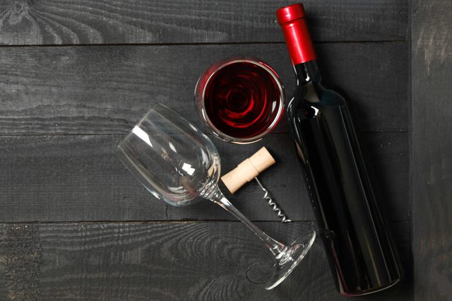 Wine bottle rests on a dark wooden table, accompanied by a corkscrew, wine glass, and filled glass beside it.