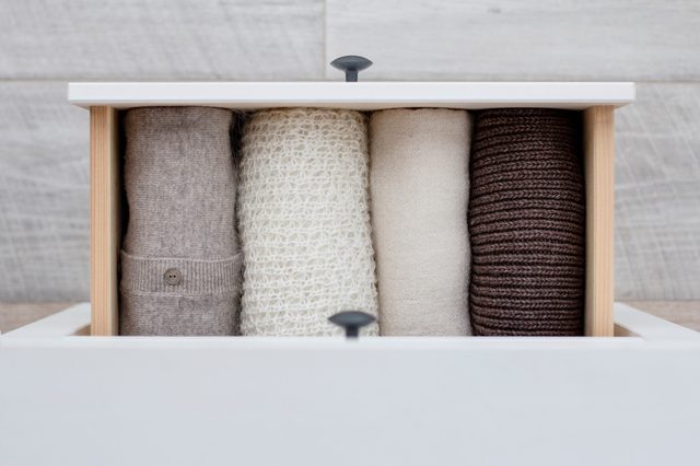 Rolled sweaters sit neatly in an open drawer, showcasing beige, white, cream, and brown colors, against a wooden background.